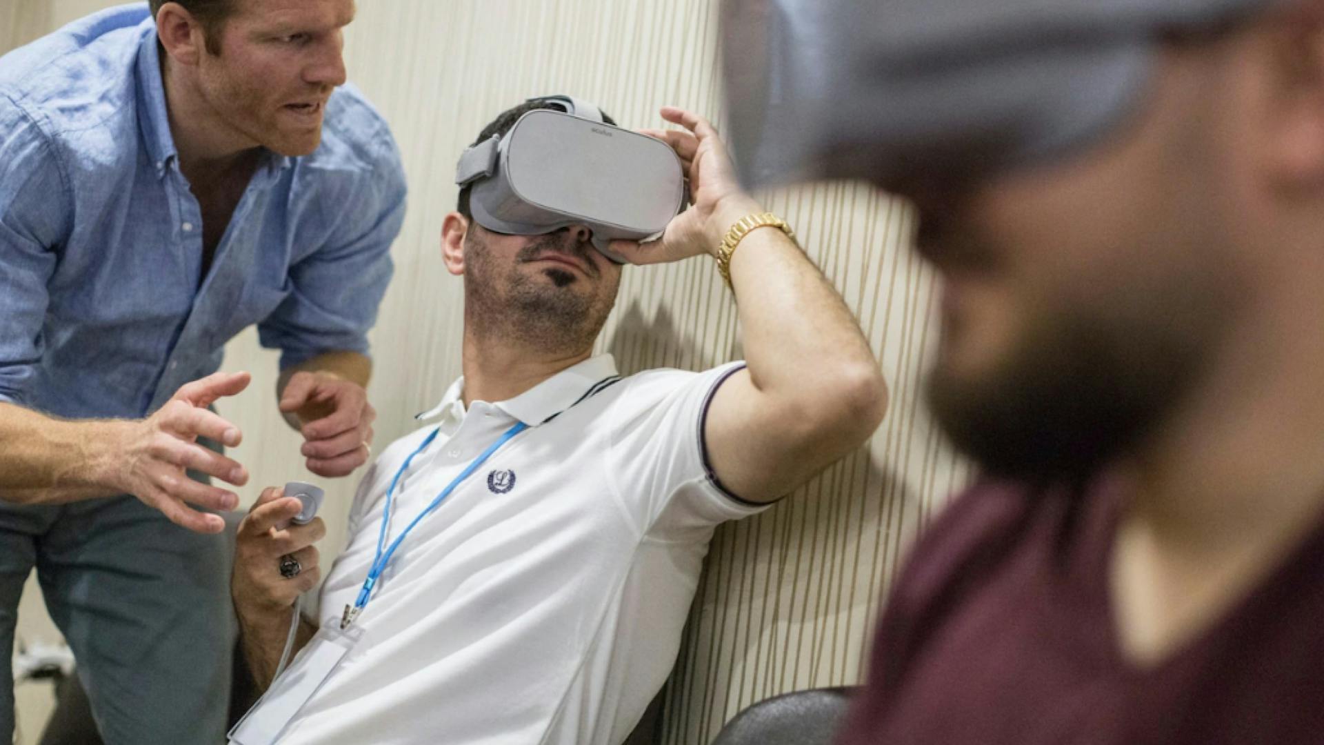 A man is sitting down, testing out the VR headset, there's a man beside him providing instructions and another man seated in the foreground, also wearing a headset.