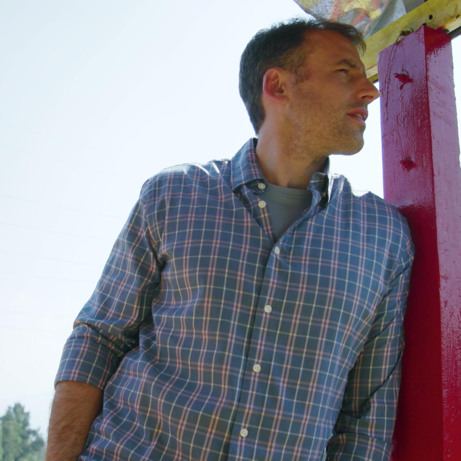 A man wearing a check shirt leads against a red post. 