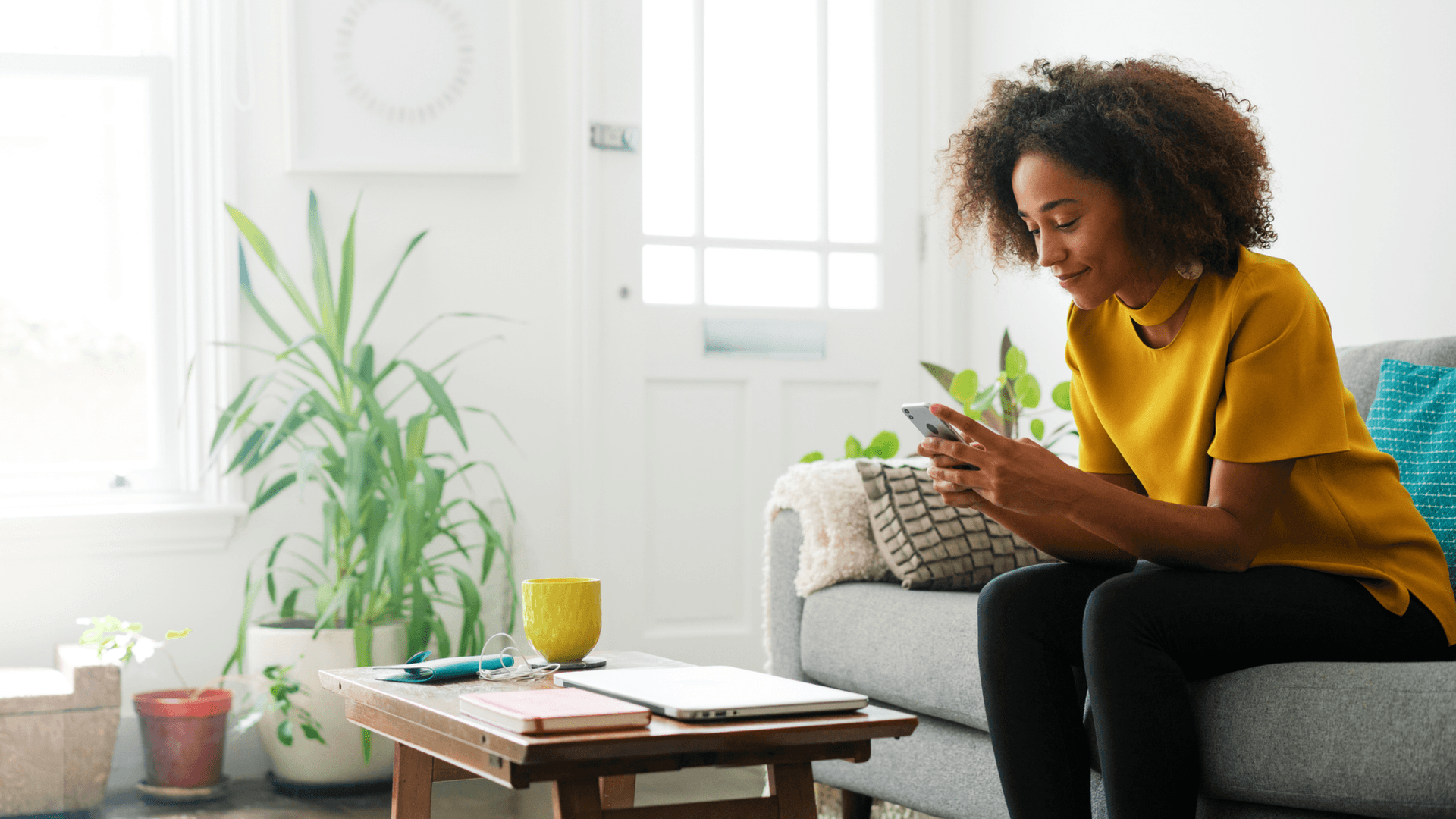 A woman sits on a sofa, smiling while looking at her phone.