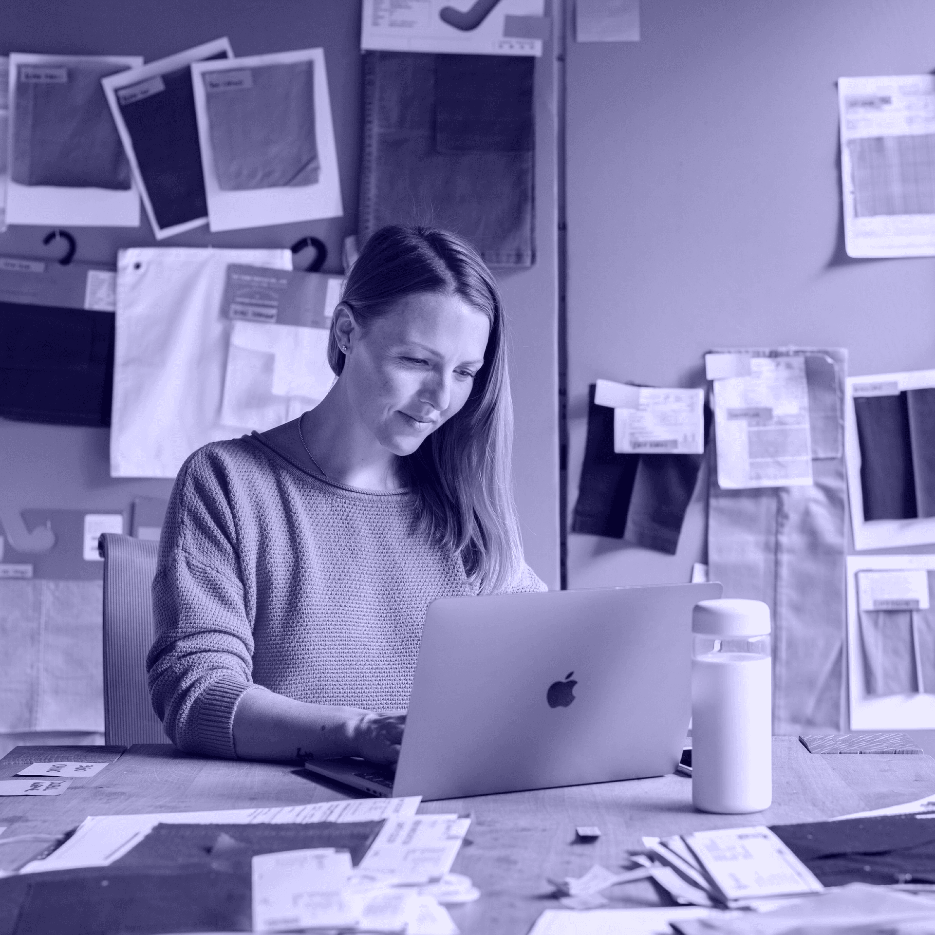 A woman sitting at a desk, looking at her laptop. Lots of papers are on the wall behind her. A woman sitting at a desk, looking at her laptop. Lots of papers are on the wall behind her.