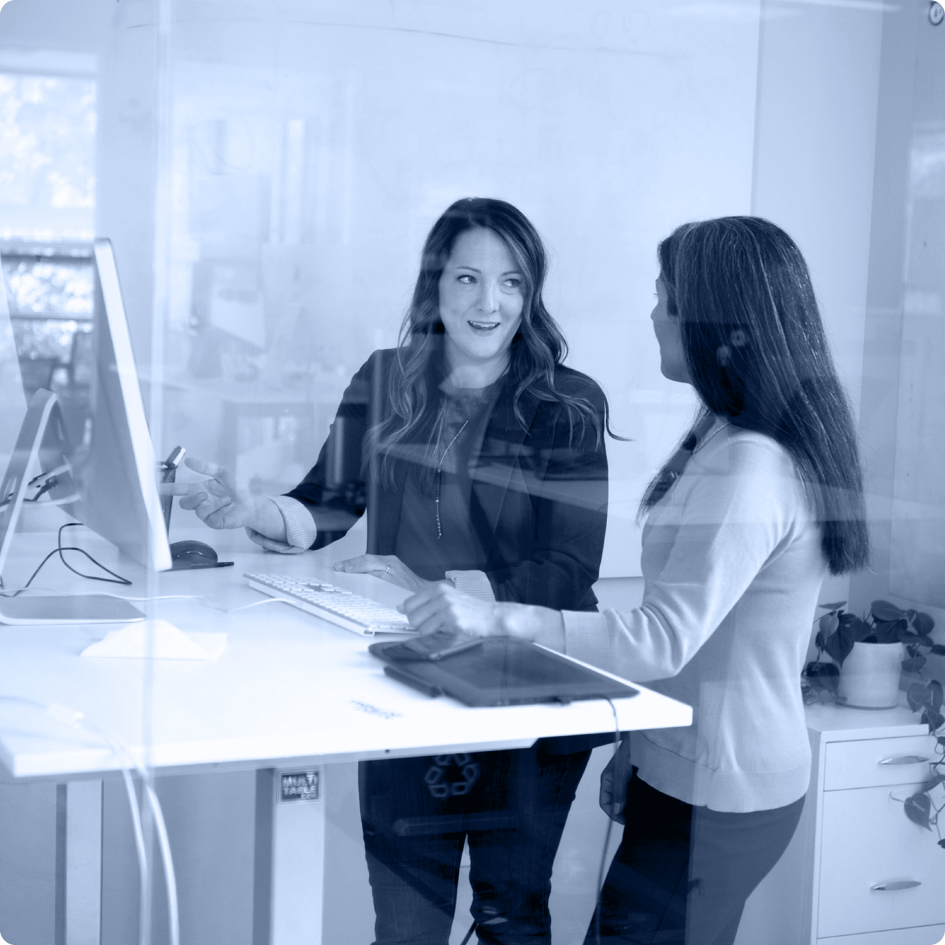 A photo of two people with long hair talking at a standing desk. A photo of two people with long hair talking at a standing desk.